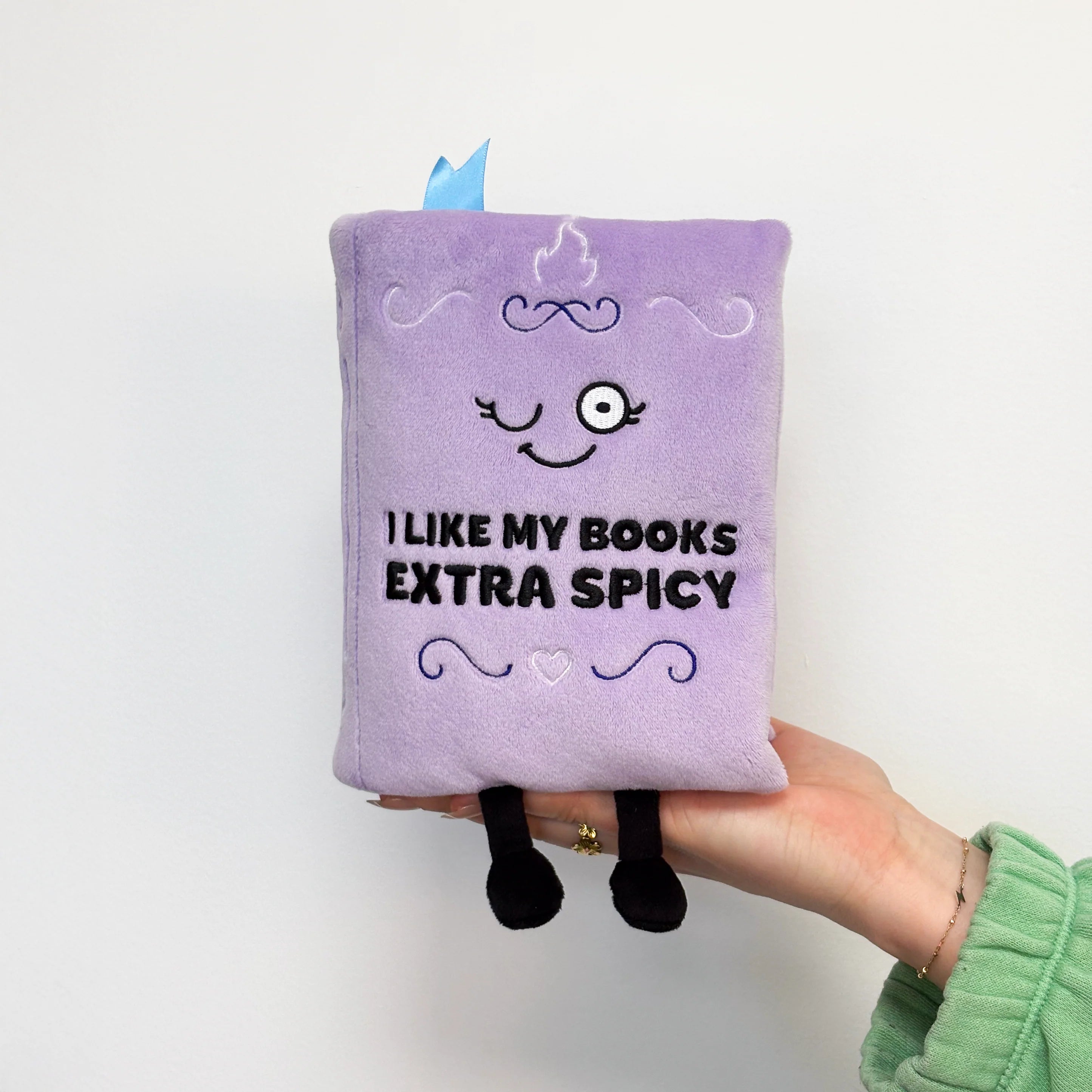 Purple plush pillow with 'I Like My Books Extra Spicy' text held by a hand on a white background